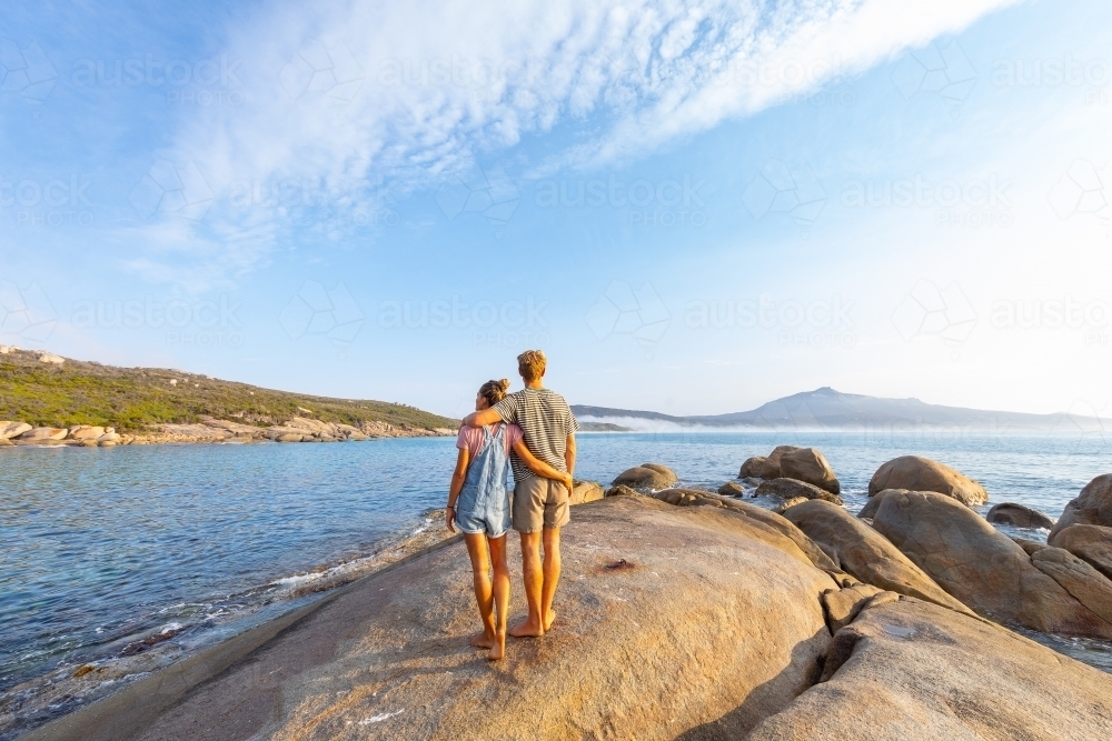 couple with arms around each other standing on granite rock looking out to sea - Australian Stock Image