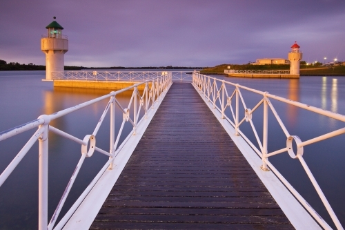 A jetty with white railings leading out to a pair of lighthouses at twilight