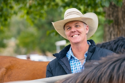 A smiling horseman wearing a cowboy hat standing among his horses