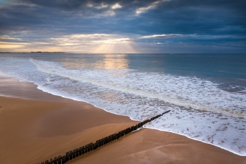 Aerial view of waves washing over a wooden groyne on a sand beach at sunrise