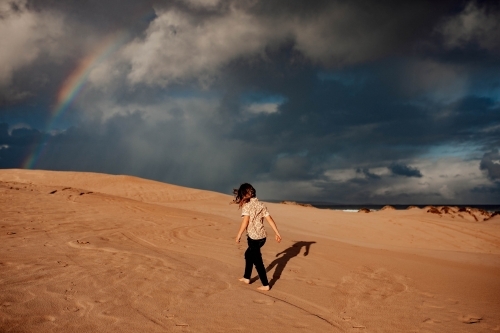 Boy walking on sand dune with rainbow in sky