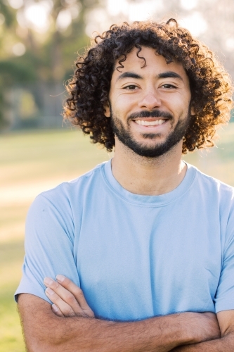 close up shot of a man smiling with curly hair and beard with crossed arms wearing blue shirt