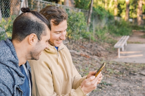close up shot of two men looking at a cellphone smiling with a white bench in the background