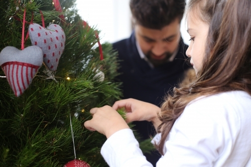 Girl decorating Christmas tree with dad in background