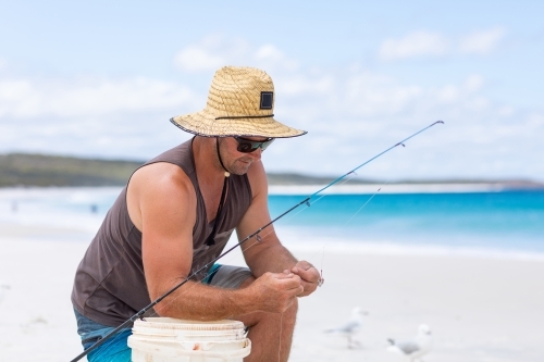 man on white sand beach kneeling down baiting fishing line