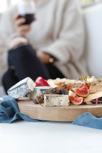 Close up of cheese platter with blurred seated woman holding wine glass in background