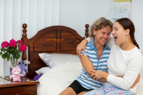 Mother and teen daughter sitting on bed and laughing with arms around each other