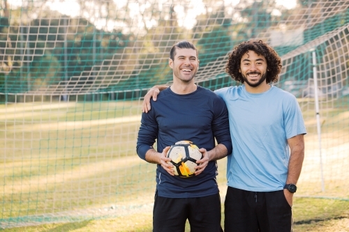 Two men smiling as they stand together in a soccer  goal, with one of the men holding a soccer ball