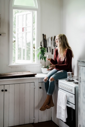 Vertical shot of a woman sitting on the kitchen counter near the window while drinking coffee