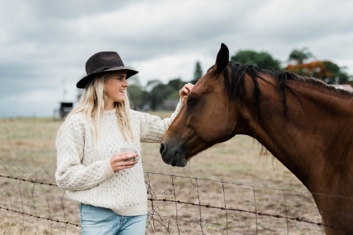 Woman holding a cup of coffee while the pats a horse