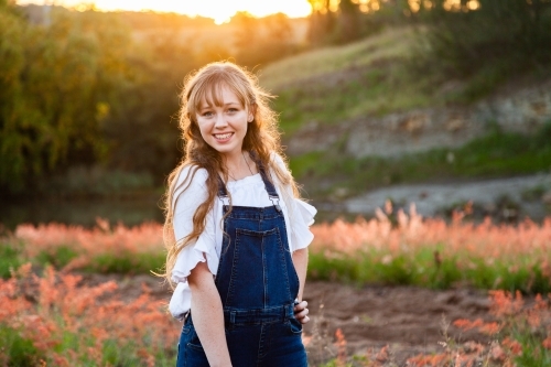 Young adult standing outside by river at sunset in overalls