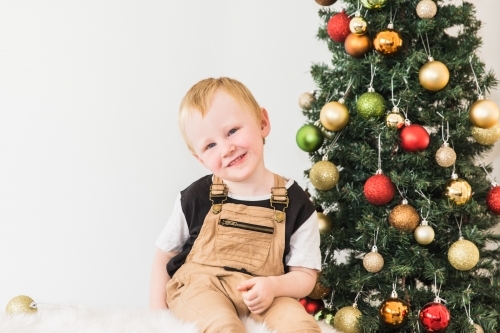 Young boy sitting next to Christmas tree with baubles smiling with head tilted