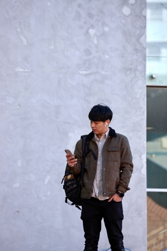 Young Chinese university student standing using mobile phone and wireless headphones