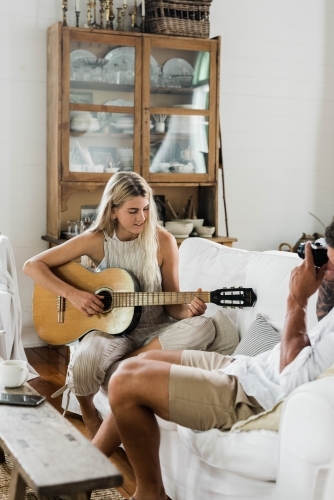 Young couple sitting on a couch, with man taking  a photo of woman playing guitar.