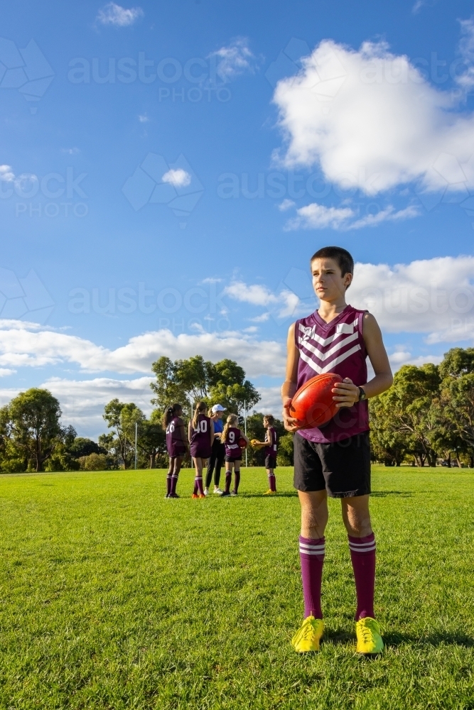 12 year old boy standing holding aussie rules football, with teammates in background - Australian Stock Image