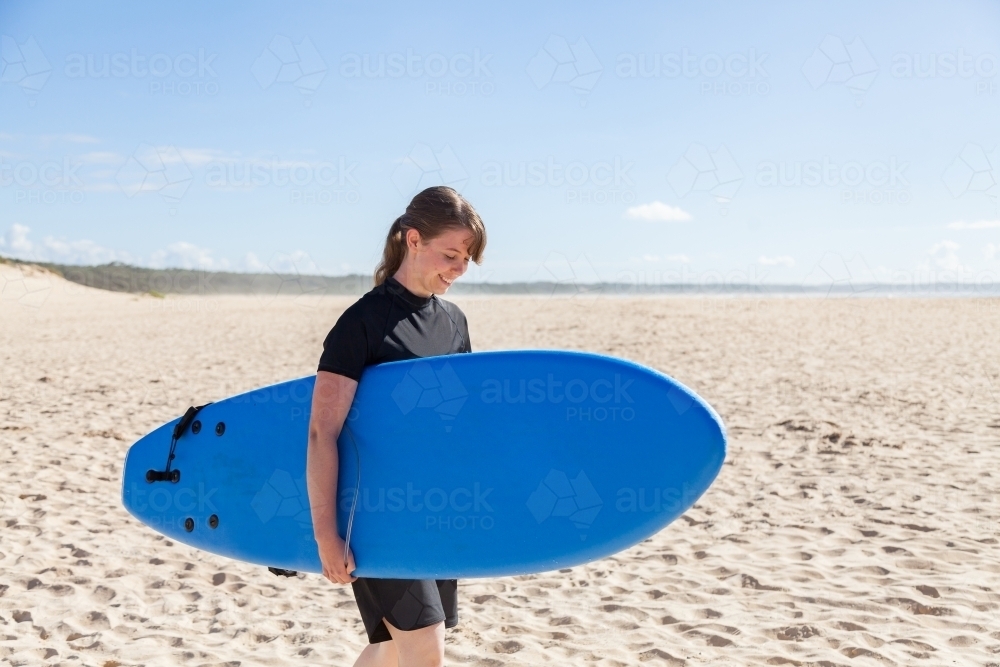 16 year old girl carrying a surfboard down the beach to the ocean - Australian Stock Image