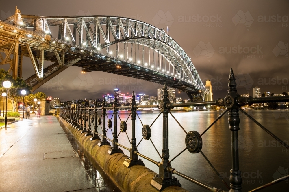 Wrought Iron fence along Sydney Harbour leading to The Sydney Harbour Bridge at night after the rain - Australian Stock Image