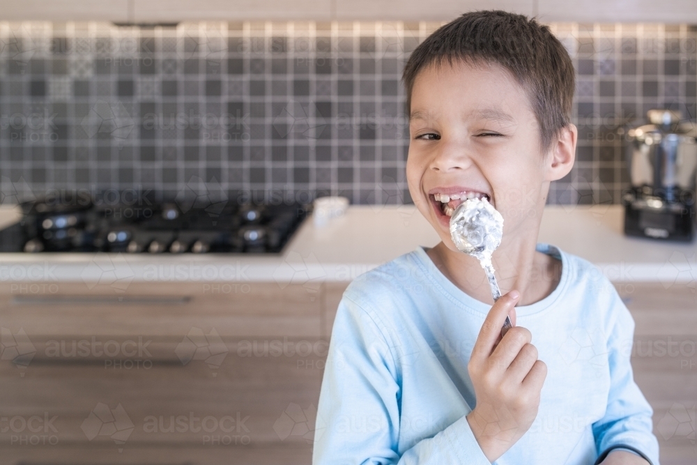 7 year old mixed race boy winks while licking a spoon of cream in his home kitchen - Australian Stock Image