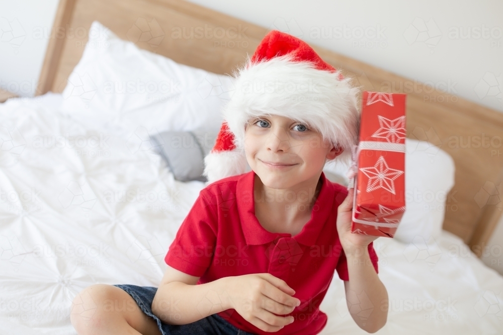 A boy wearing a red shirt sitting on a bed wearing a Santa hat shaking a wrapped christmas present, - Australian Stock Image