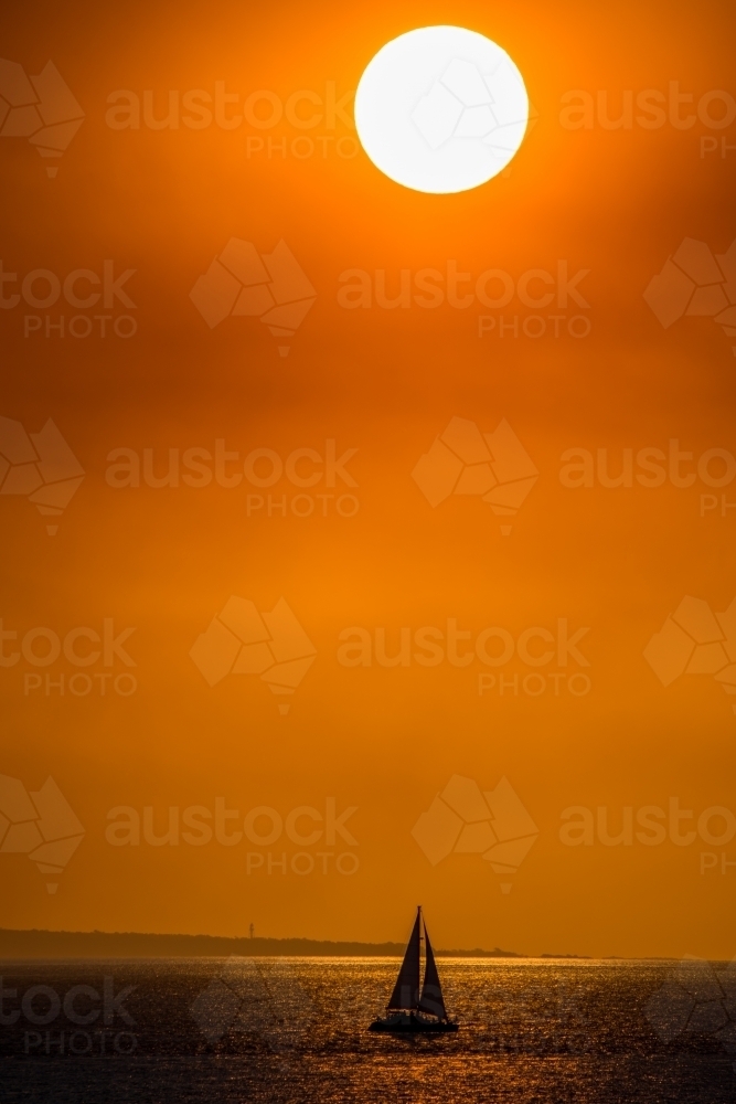 A bright sun hangs low in the sky with a small silhouette of a sailboat on the ocean. - Australian Stock Image