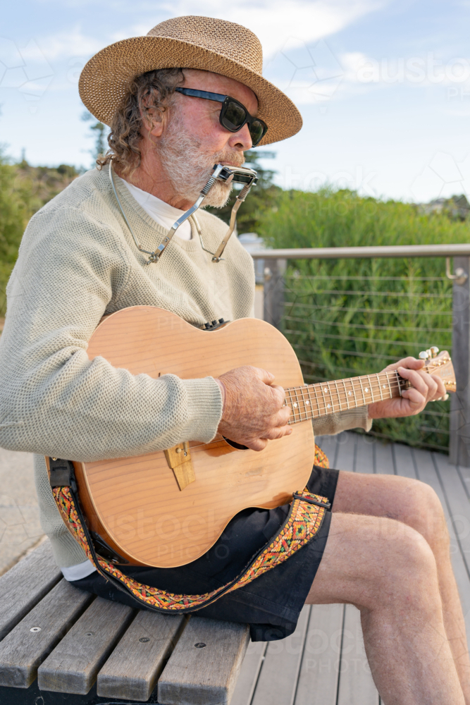 A busker wearing a hat playing a guitar and harmonica on a park bench - Australian Stock Image