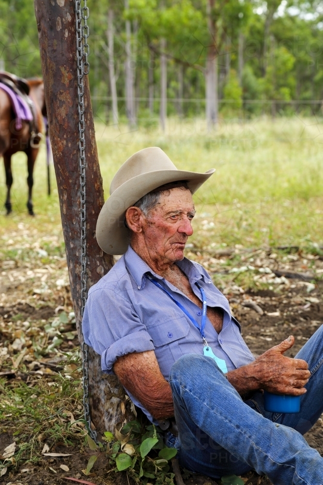 A cattle drover in his eighties rests against a tree with a cup of tea - Australian Stock Image