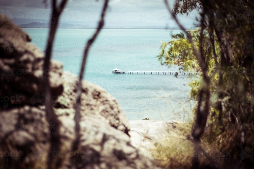 A coastal view with a long pier or jetty that can be seen through rocky formations and tree branches - Australian Stock Image