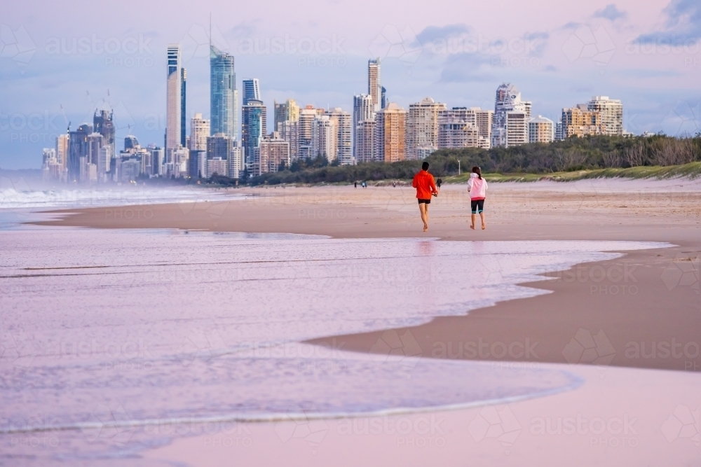 A couple running along a beach at twilight in front of hi rise buildings - Australian Stock Image