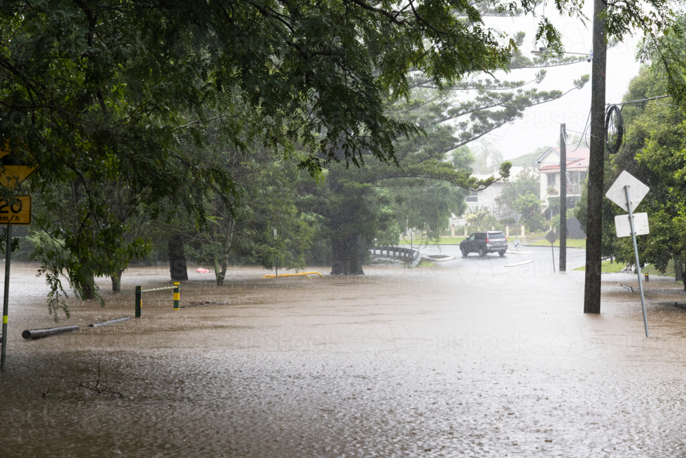 A flooded road and park in a low lying area of Red Hill. A car drives away from floodwater in rain - Australian Stock Image