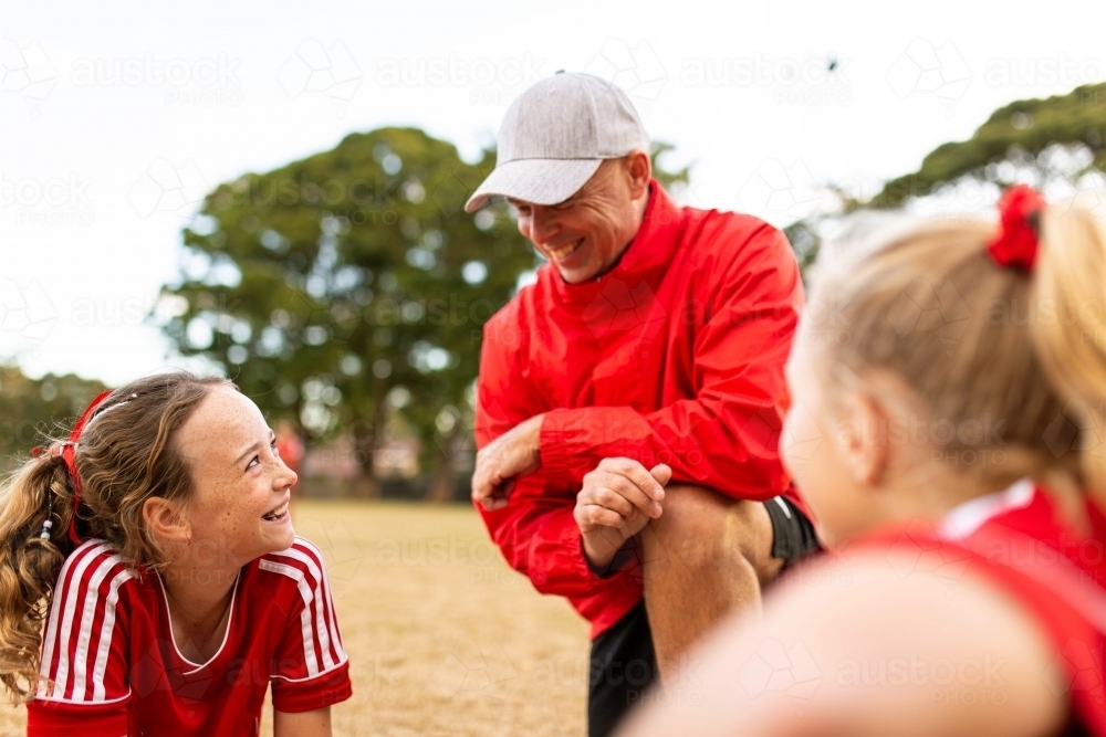 A football team coach kneeling down talking to young girls in the football team at the field - Australian Stock Image