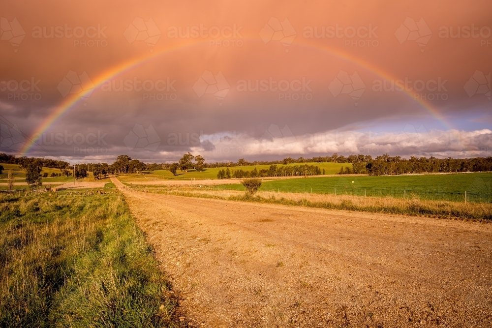 A ground level view looking down a dirt road with a vivid rainbow arching overhead - Australian Stock Image