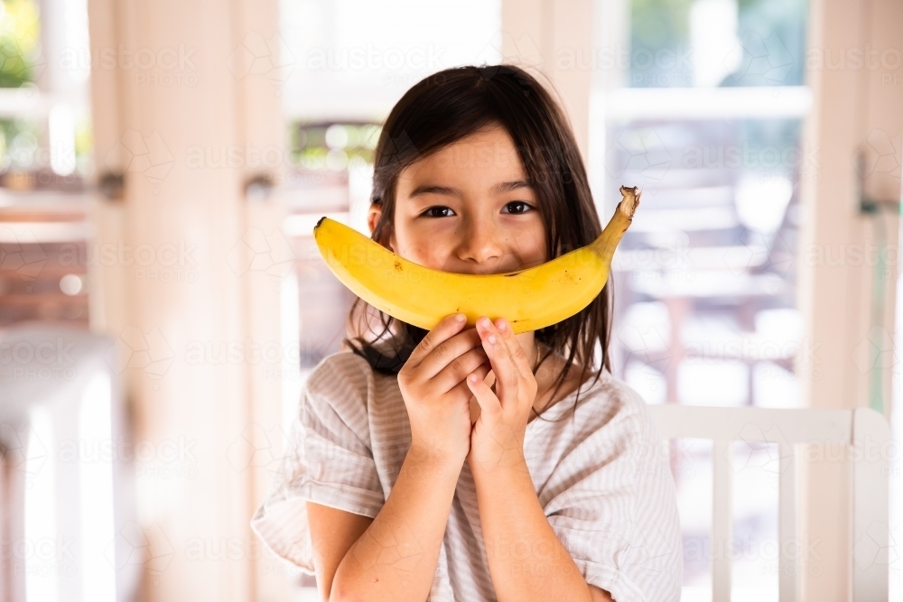 A happy little girl holding a big banana in the kitchen at home - Australian Stock Image