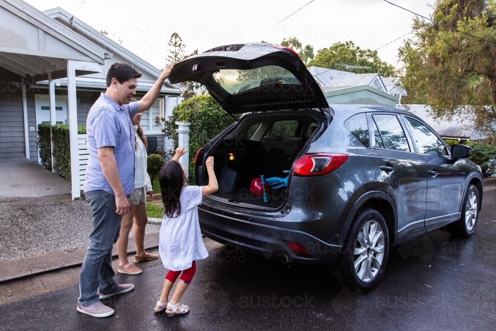 A little girl in white dress helping dad close the car boot ready to go to park - Australian Stock Image