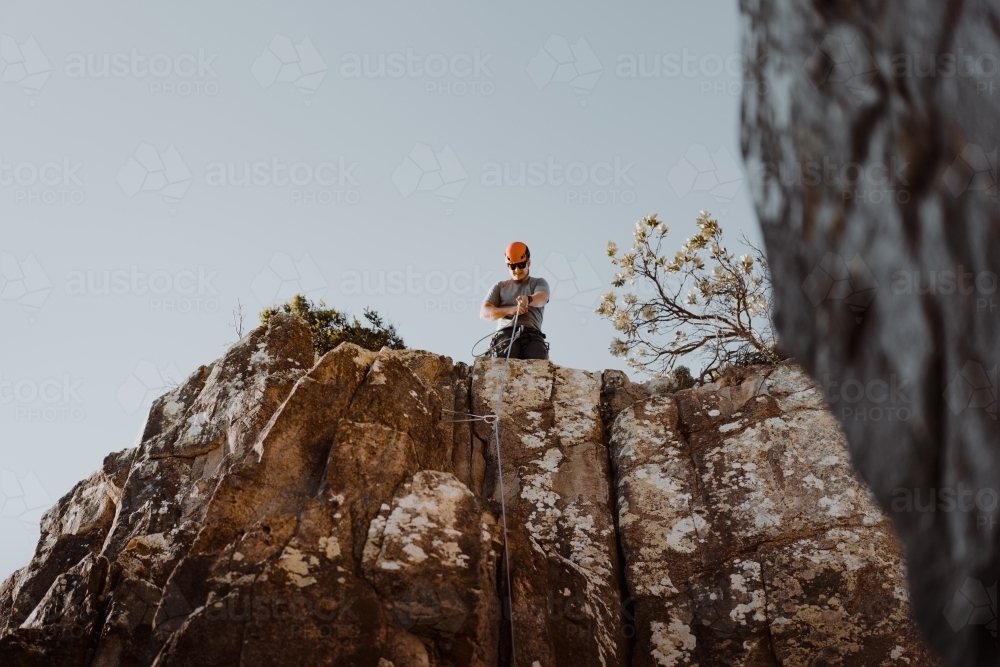 A male rock climber pulling rope through at the summit of a climbing route. - Australian Stock Image