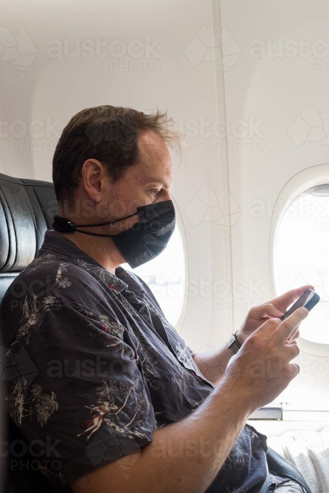 A man wearing a mask and extender while looking at his phone on an airplane - Australian Stock Image