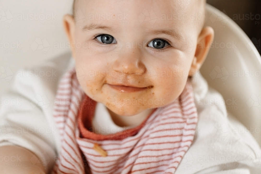 A one year old baby with blue eyes, she has a dirty face as she has been feeding herself spaghetti - Australian Stock Image