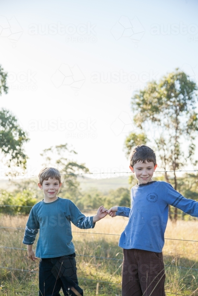 A pair of boys leaning against a barbed wire fence in paddock outside - Australian Stock Image