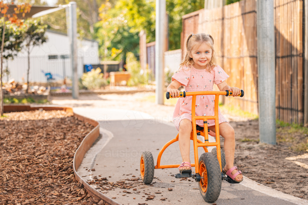 A preschool age girl riding an orange trike at kindergarten - Australian Stock Image