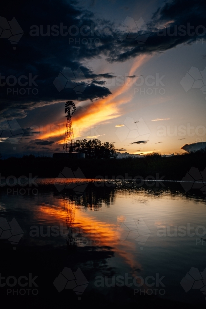 A silhouette of the tall windmill and trees at sunset by dam - Australian Stock Image