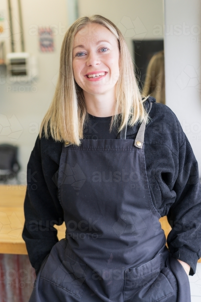 A smiling young woman barber with her hands in her pockets and wearing  an apron - Australian Stock Image