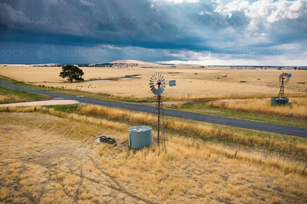 A windmill on the roadside of dry rural farmland with dark clouds and rain falling in the distance - Australian Stock Image