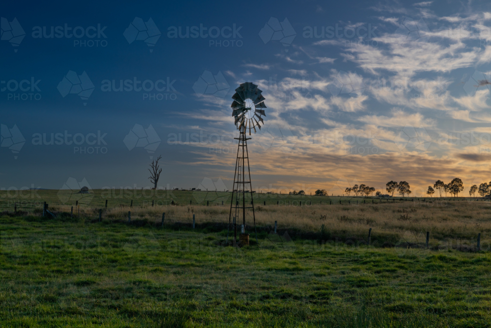 A wispy cloudy sky and a windmill with a silhouette of a tree trunk on an early morning - Australian Stock Image