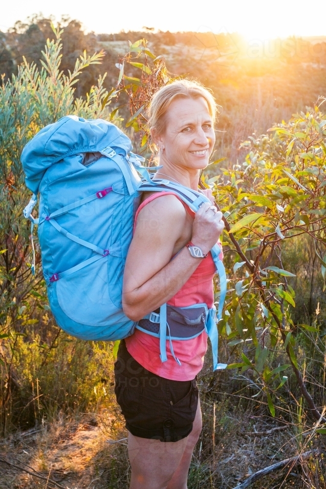 A woman hiker carrying a backpack looking back over her shoulder at sunset - Australian Stock Image