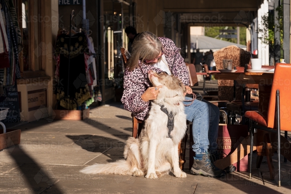 A woman seated at an outdoor cafe table, patting her pet dog - Australian Stock Image