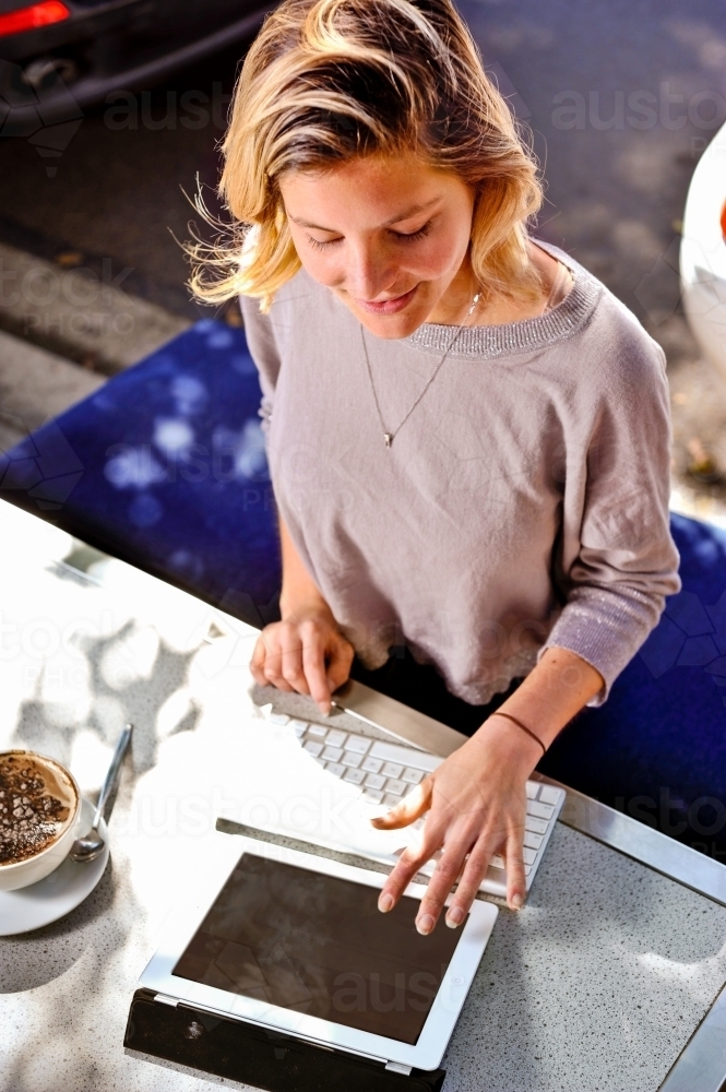 a young woman enjoys her cappuccino whilst working outdoors in a cafe with her tablet device - Australian Stock Image