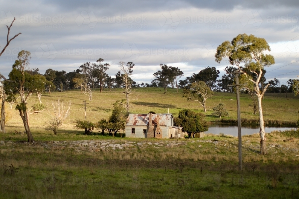 Abandoned workmans hut situated in a paddock by a dam full of water caught in the afternoon sun - Australian Stock Image