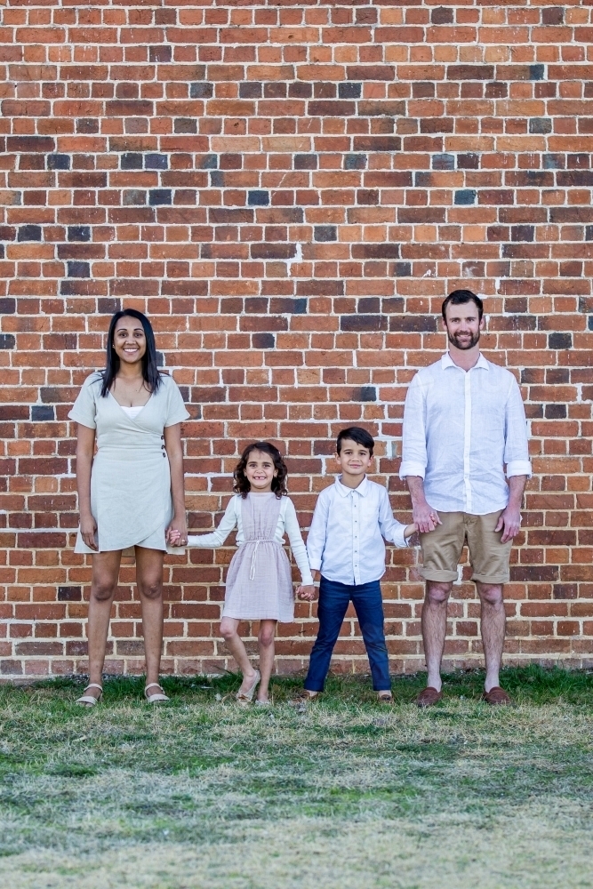 Aboriginal and Caucasian mixed race family standing against brick wall holding hands - Australian Stock Image