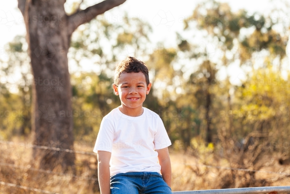 Aboriginal Australian boy sitting on farm gate in paddock smiling - Australian Stock Image