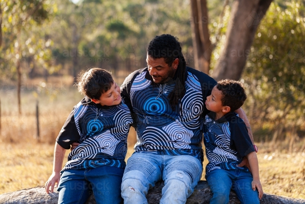 Aboriginal dad sitting together with sons on log in bush - Australian Stock Image