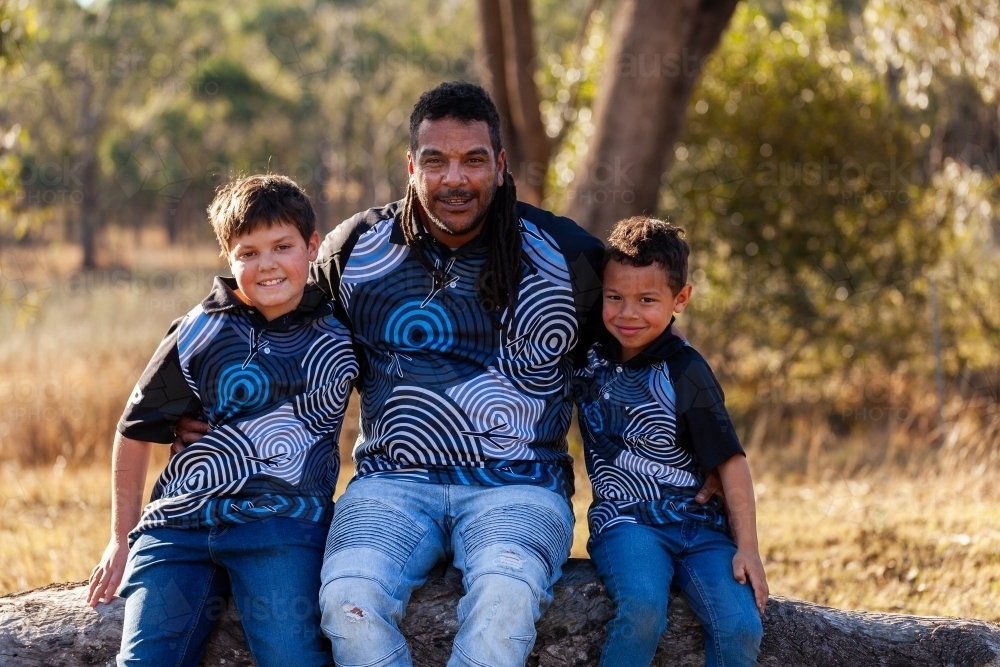 Aboriginal dad sitting together with sons on log in bush - Australian Stock Image